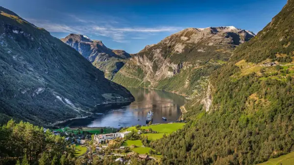 Ein Kreuzfahrtschiff gleitet durch die majestätischen norwegischen Fjorde, umgeben von steilen, bewaldeten Bergen und einem kleinen Dorf.